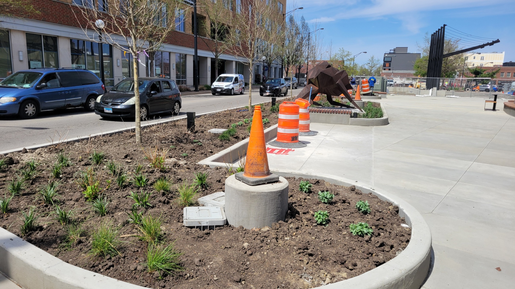 A view of a concrete pedestal with a traffic cone on top where a light pole will be installed. In the background, two traffic barrels denote where the bench swing structure will be installed next to the public art sculpture known as "strider."