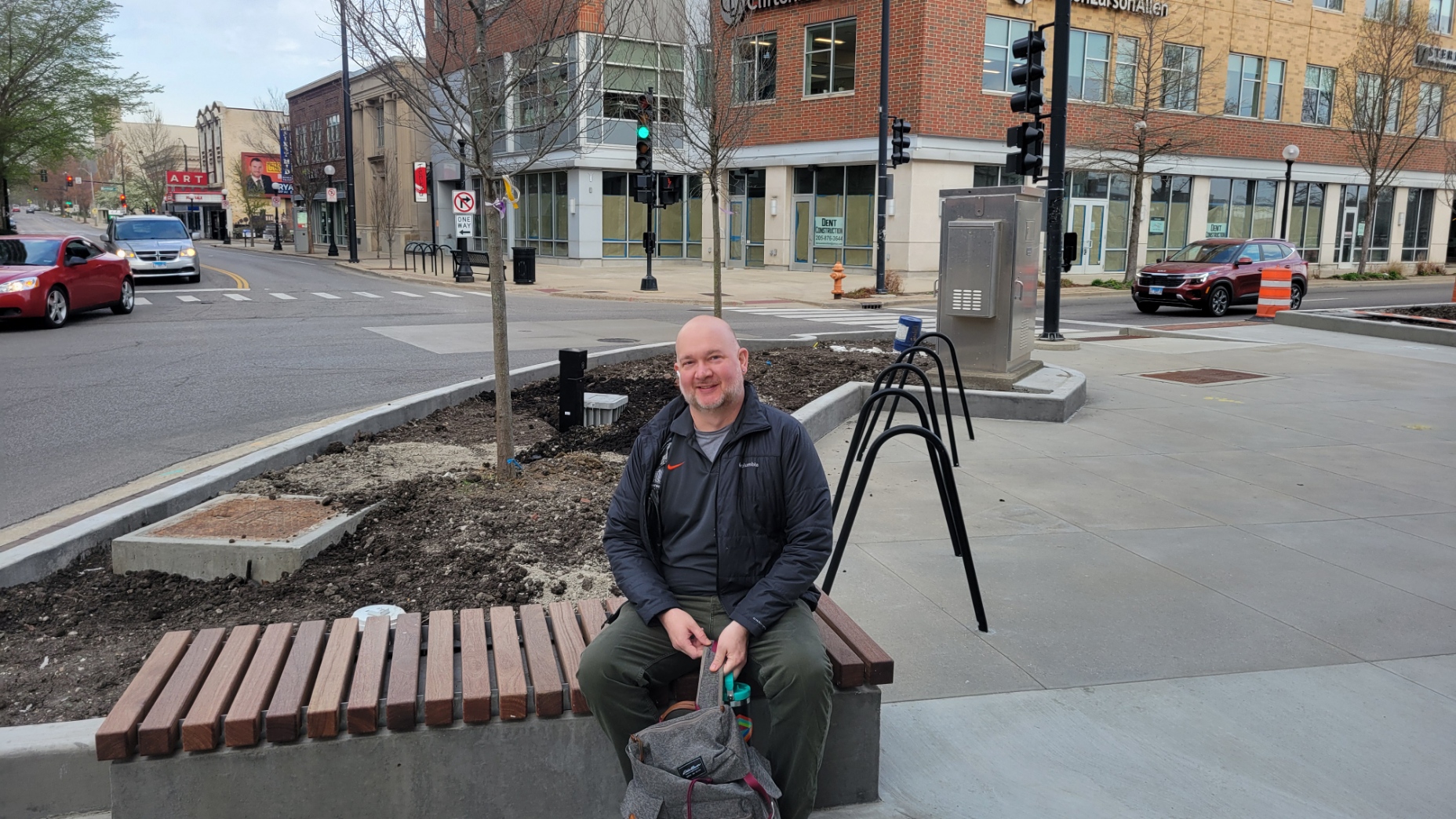 Gentleman sits on a woodcapped bench on the south side of Phase 1 by bike racks and a planter near the intersection of Main and Neil