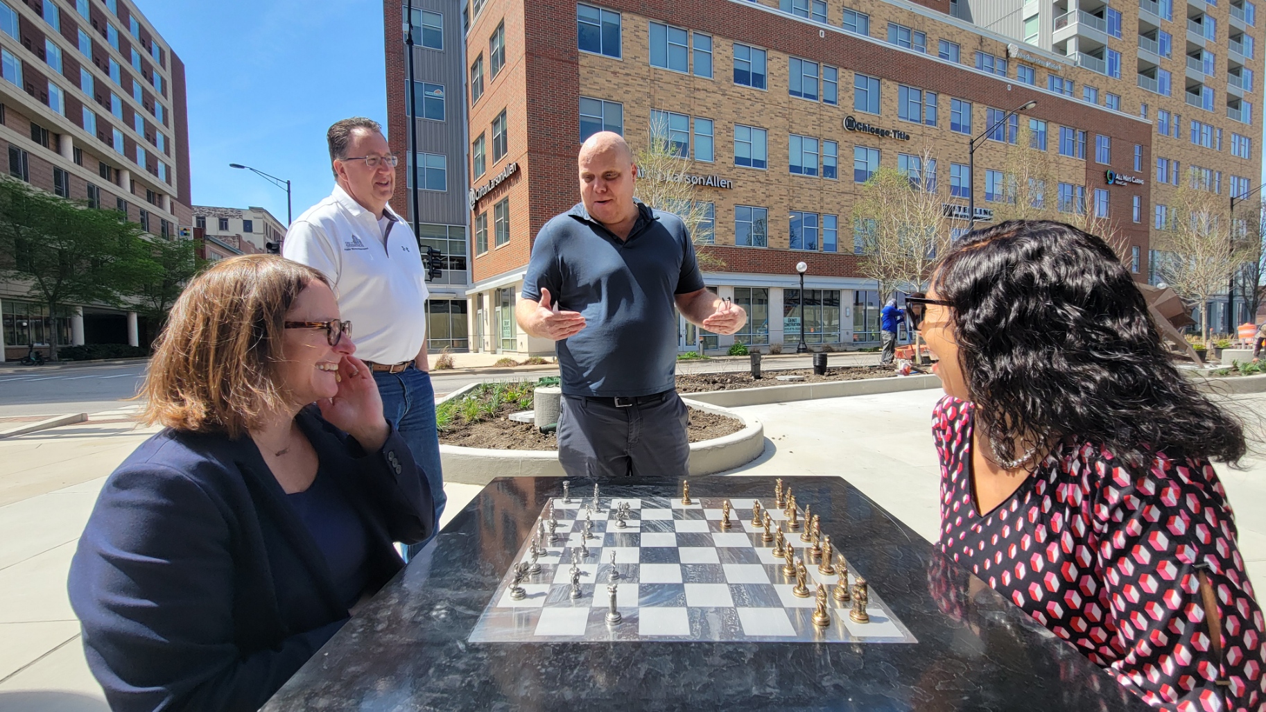 Mayor Deb Feinen and Joan Walls smile over chess board in the plaza, listening to Scot McIntosh (nearby restaurant manager) in the background. Also featured is project engineer David Happ.