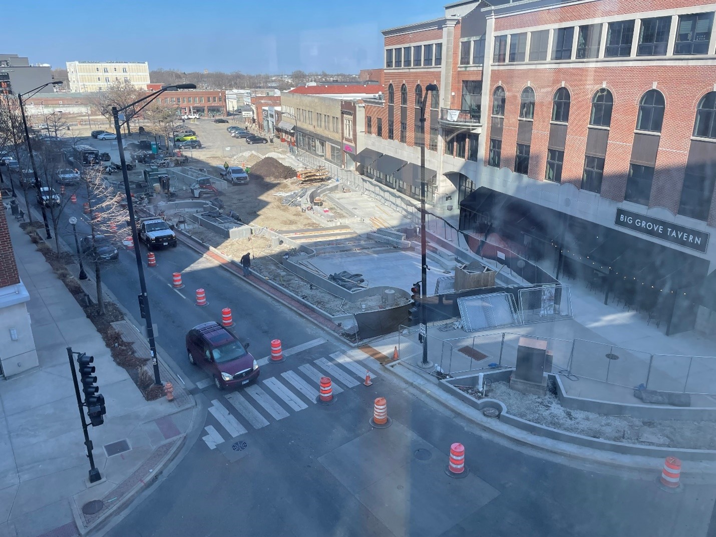 A bird's eye view photo of the plaza construction work progress as of early March 2025, facing northeast. Concrete for planters has been poured, about one third of concrete for the walking surface has been poured, and the foundation for the stage is in progress. Construction materials, workers, and vehicles are present.