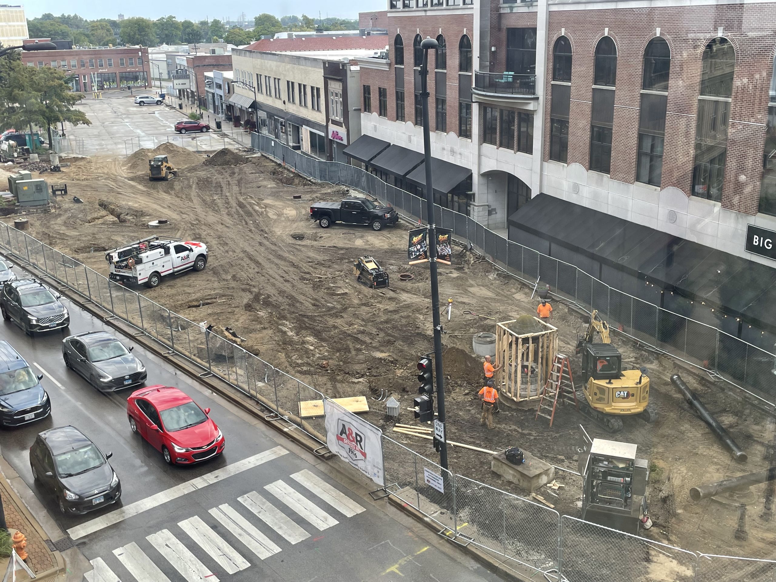 A photo taken from the fourth floor of a parking deck southwest of the project, facing northeast. The current project area is shown with construction workers, construction equipment, a protective wooden structure around an historic well and construction fencing surrounding the active work area which is covered in soil.