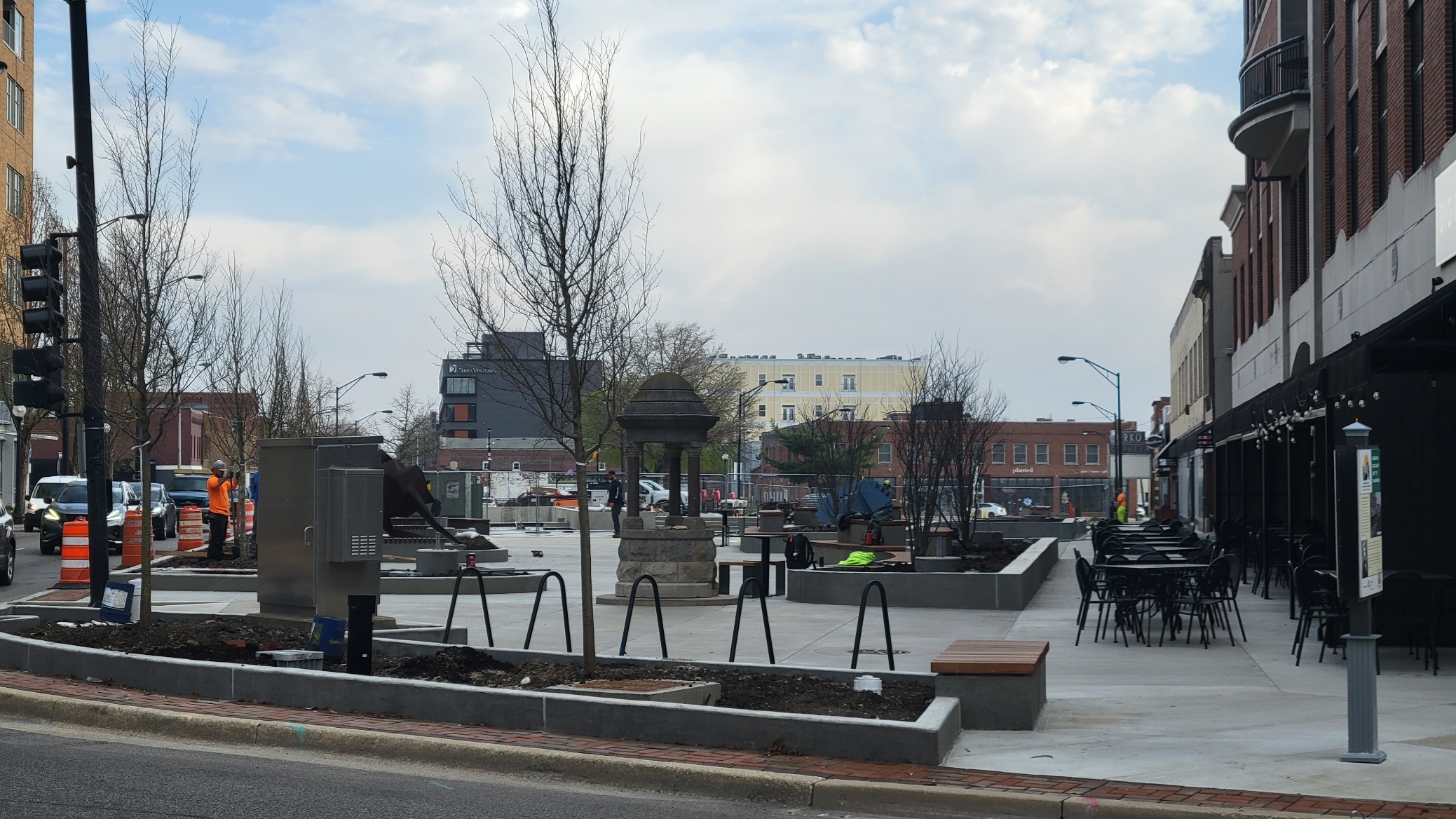 A view of the downtown plaza facing north from Main Street, featuring trees, bike loops, a historic well, and outdoor seating for a nearby restaurant.