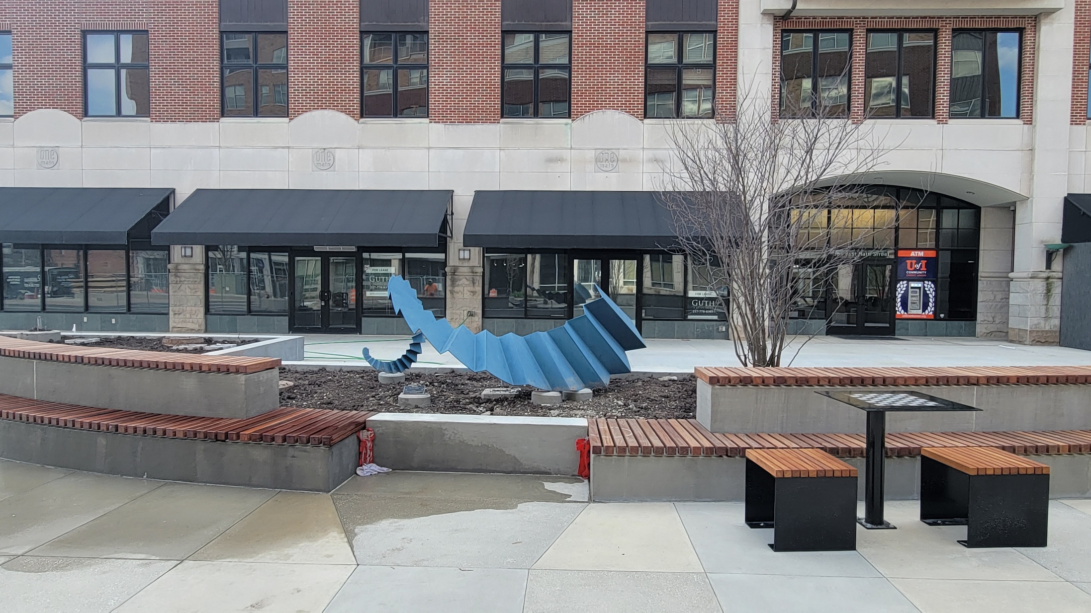 A view of tiered bench seating facing east. A public art sculpture is featured next to a tree in a landscaping planter. Also pictured are two seats and a chess table fixed to the plaza.