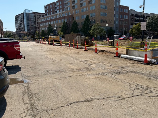 A photo of the Neil and Washington parking lot, facing south, featuring an open aisle of parking spaces adjacent to cones and construction equipment where a water main was installed.