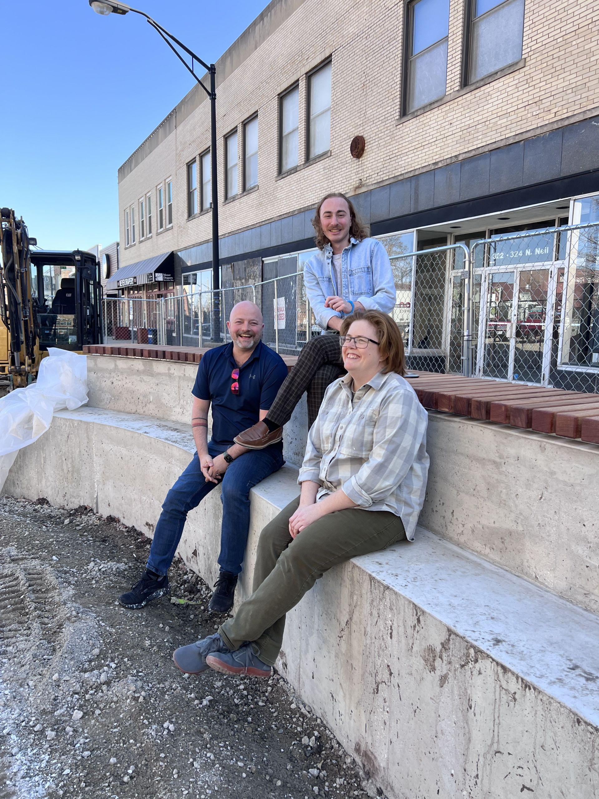 Three individuals sit on tiered bench seating that will be incorporated into landscaping planters throughout the plaza. These bench seats are concrete and feature a lower tier for seating and a higher tier for seating. The second (highest) tier has wood caps for comfortable seating.