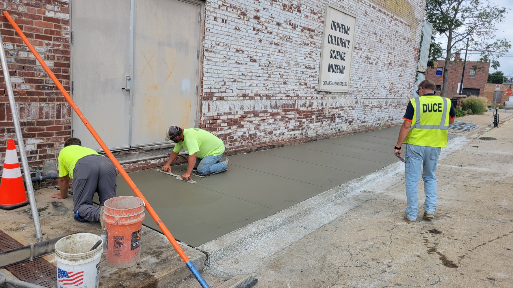 A photo of a repaired concrete sidewalk along the east side of the Orpheum Theater near the Downtown Plaza site, with construction workers and concrete still drying